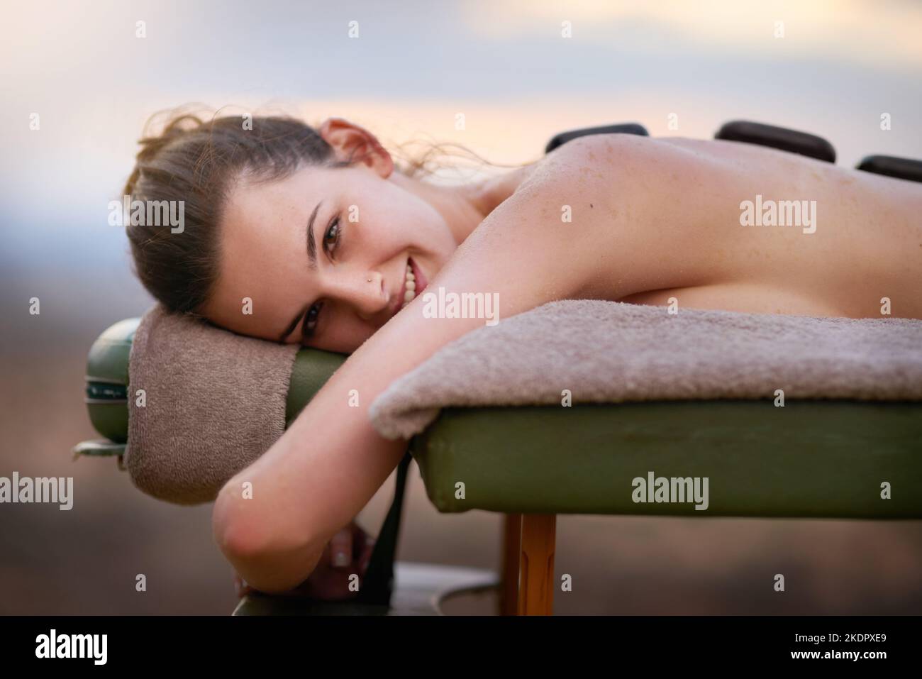 Sei gut zu dir selbst. Porträt einer jungen Frau, die draußen auf einem Massageliege eine Behandlung mit heißen Steinen erhält. Stockfoto