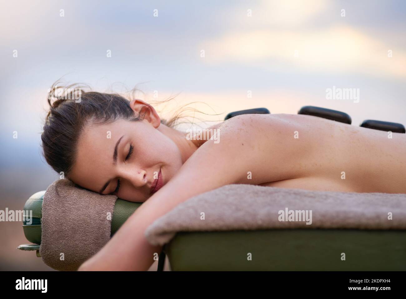 Eine junge Frau, die draußen auf einem Massageliegen eine Behandlung mit heißen Steinen erhält. Stockfoto