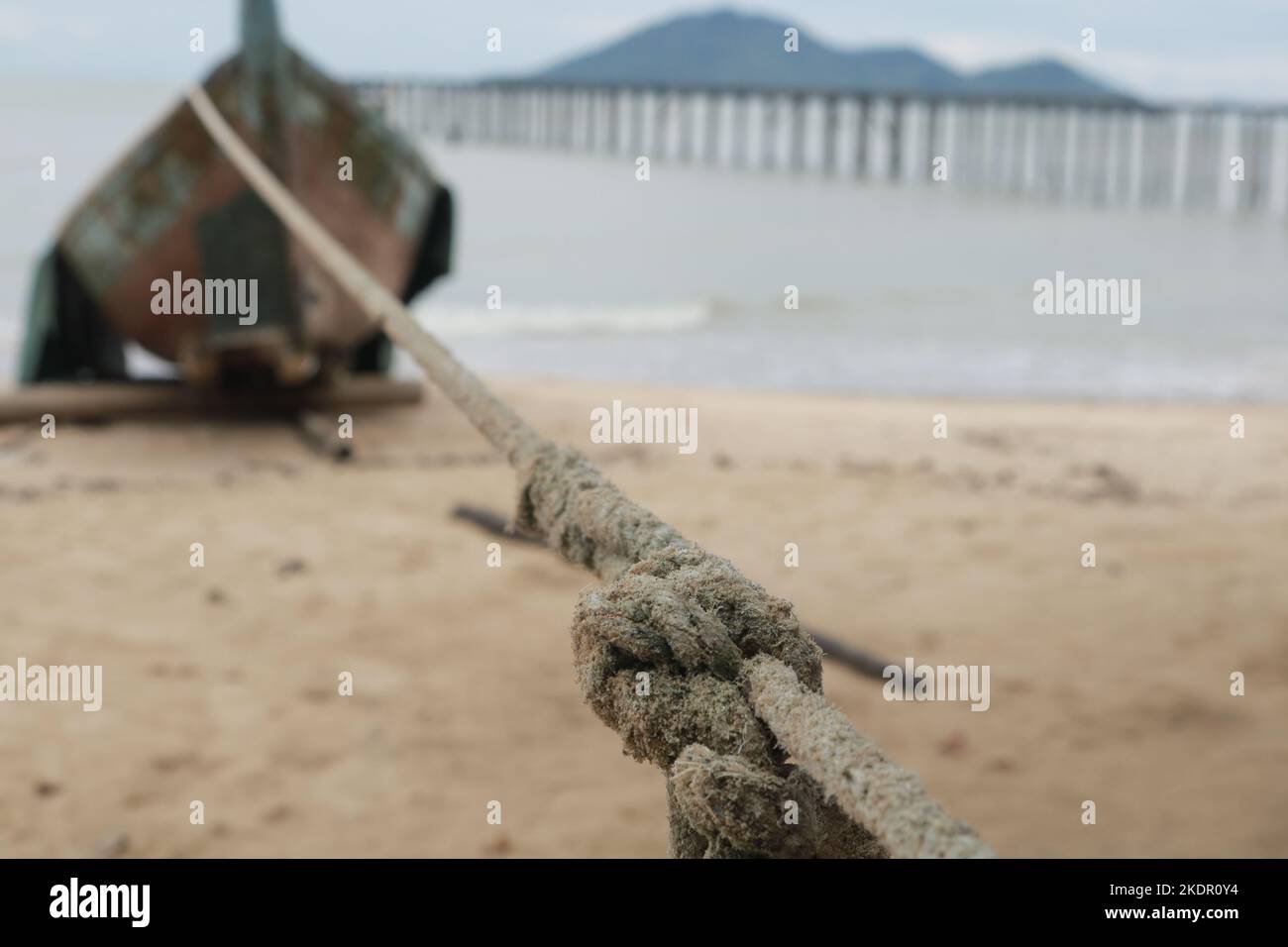 Braunes Seil mit Knoten auf Seil Krawatte verbunden mit Holzboot am Strand mit Blick auf den Strand, hölzernen Pier mit Insel und bewölktem Himmel Stockfoto