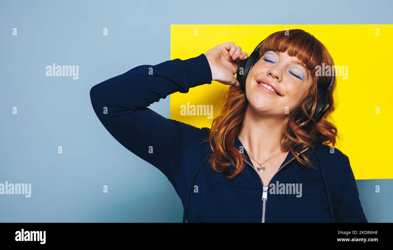 Fröhliche Frau, die in einem Studio Musik über kabellose Kopfhörer hört. Frau, die gegen eine geometrische Form steht. Stockfoto