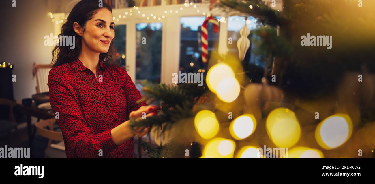 Frau, die Dekorationen auf den Weihnachtsbaum legt. Frau, die den Weihnachtsbaum in einem gemütlichen Familienhaus schmückt. Stockfoto