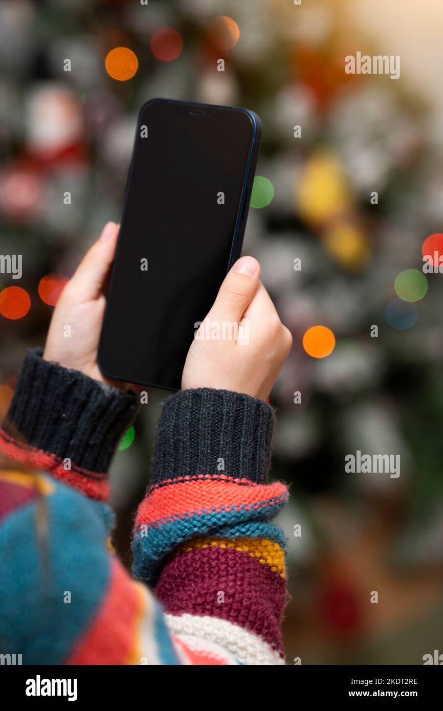 Kind hält ein leeres Smartphone mit dem Weihnachtsbaum im Hintergrund Stockfoto