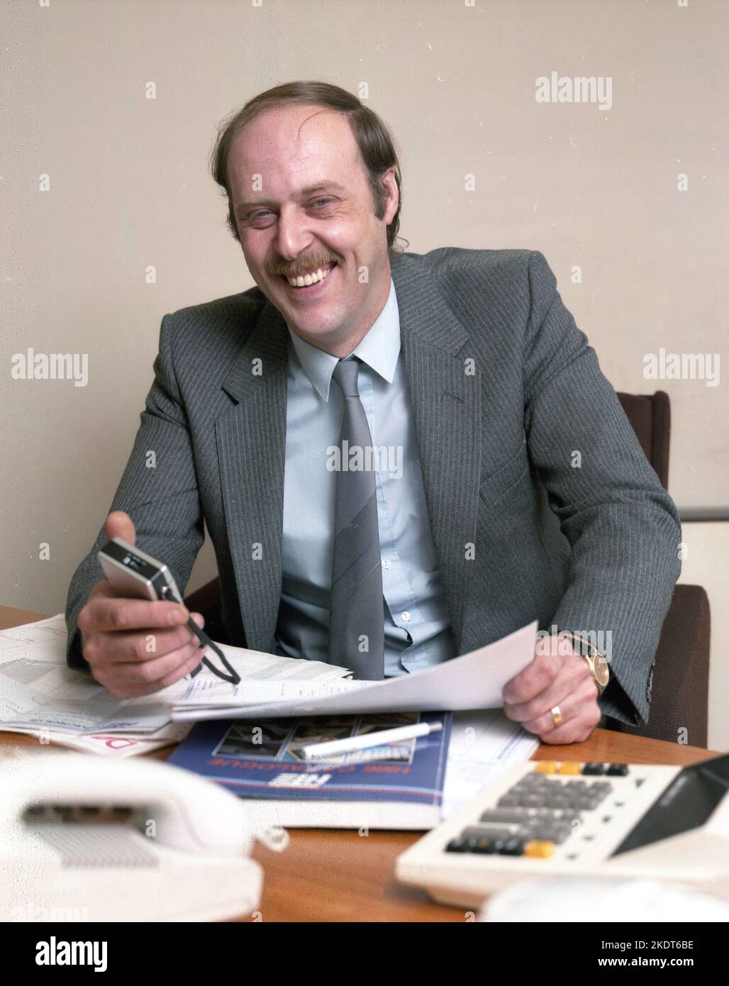 1980s, historisch, ein Geschäftsführer, der an seinem Schreibtisch sitzt, mit Papierkram und in der Hand, eine Diktatmaschine, England, Großbritannien. Stockfoto