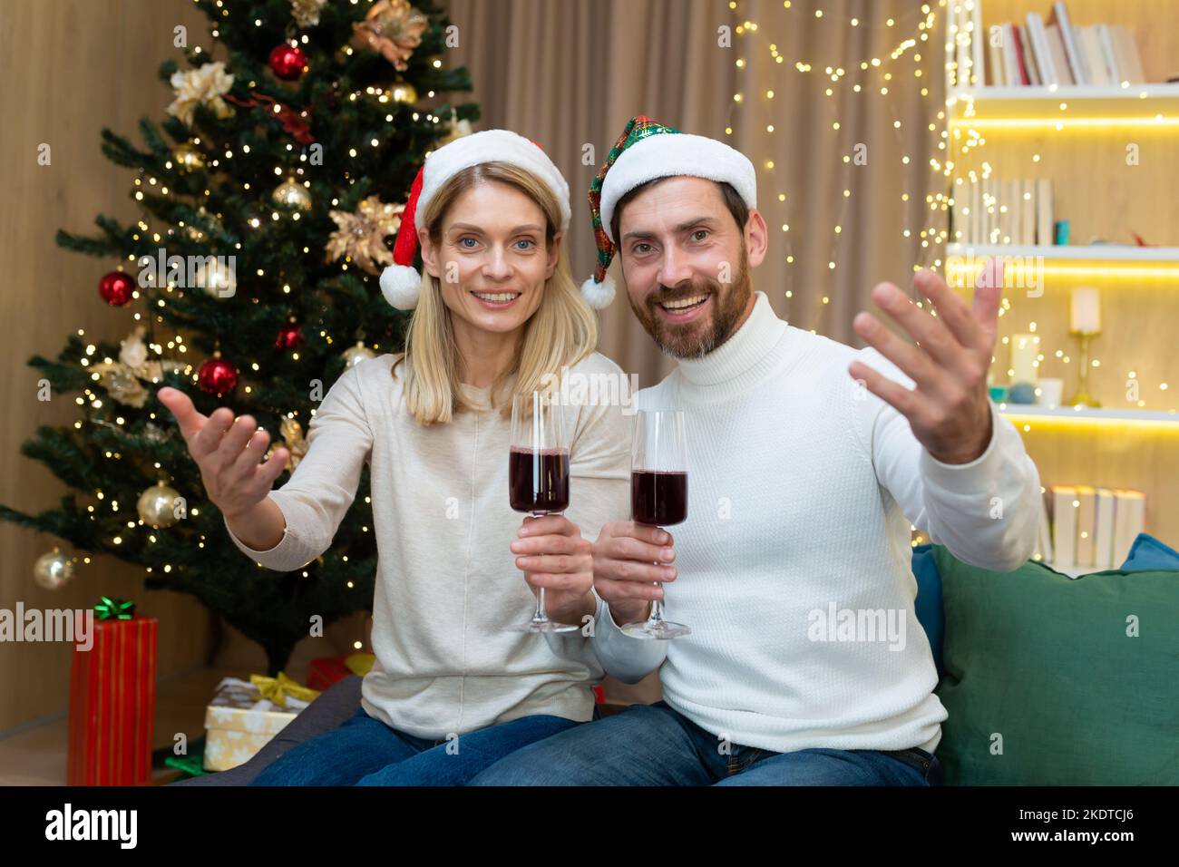 Festliches Neujahrsbild eines jungen Paares. Sie sitzen auf dem Sofa neben dem Weihnachtsbaum in roten Weihnachtsmannmützen, halten Weingläser in den Händen und laden die Gäste mit Handgesten ein. Stockfoto