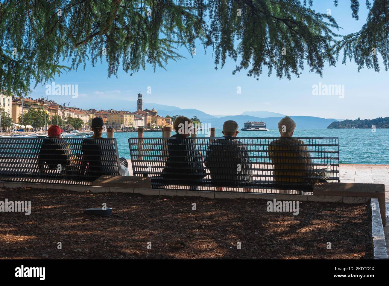 Ältere Menschen im Sommer, Rückansicht einer Gruppe älterer Menschen, die auf einer Bank sitzen und die malerische Stadt am Gardasee in Salo, Lombardei, Italien, betrachten Stockfoto