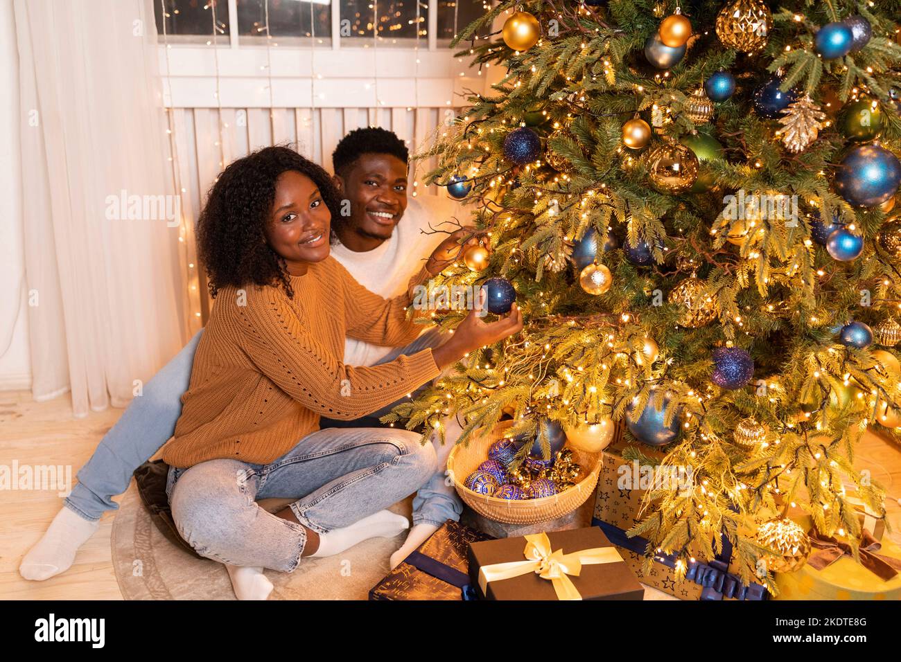 Die junge afroamerikanische Dame und der junge Mann schmücken den Weihnachtsbaum mit Girlanden, Spielzeug im gemütlichen Wohnzimmer Stockfoto