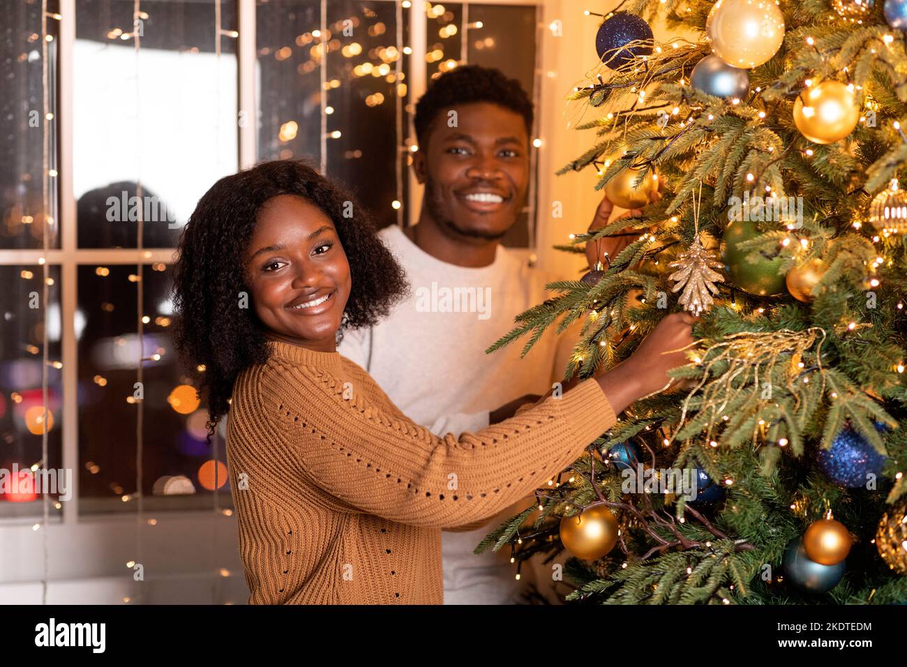 Glückliche tausendjährige afroamerikanische Frau und Mann schmücken Weihnachtsbaum mit Girlanden, Lichter Stockfoto