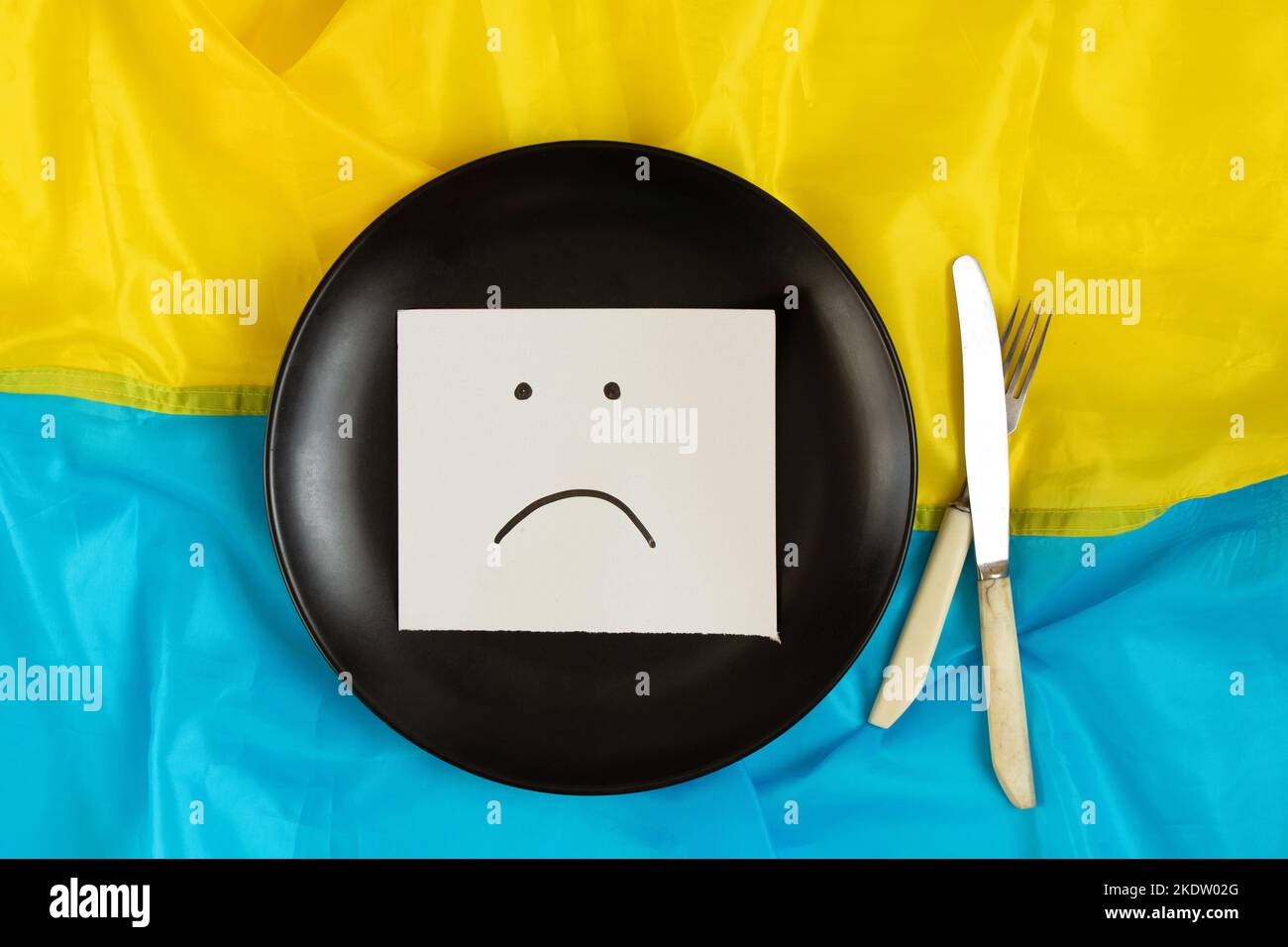 Ein trauriges, auf Papier gezeichnetes Emoticon liegt auf einer schwarzen Platte auf den Flaggen der Ukraine, schlechte Laune, kein Essen und Hunger aufgrund des Krieges 2022 Stockfoto