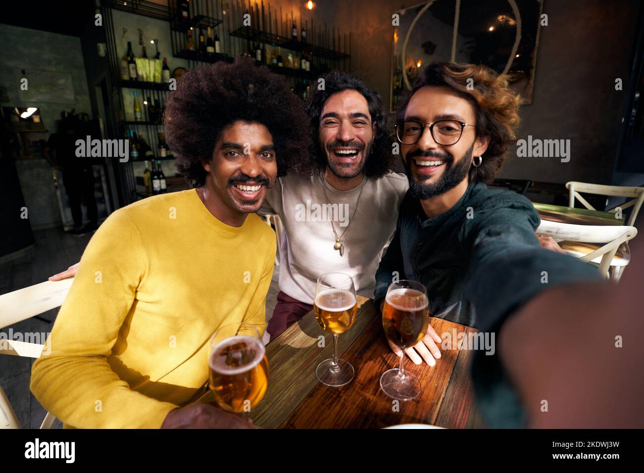 Horizontales Selfie von drei männlichen Freunden, die fröhlich in einer Bar toasten und mit großem Lächeln auf die Kamera blicken. Stockfoto