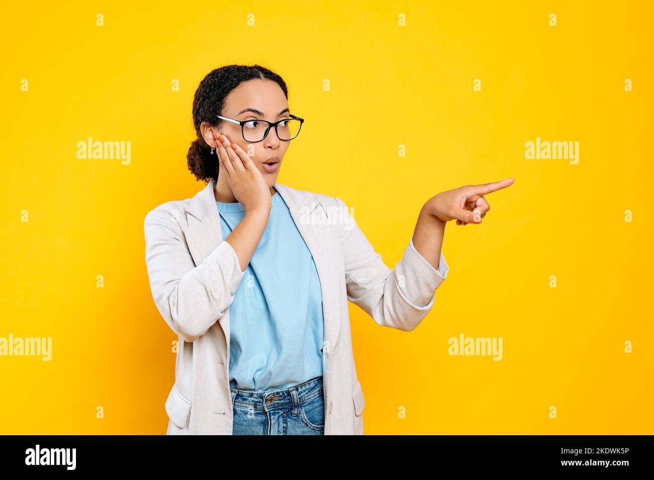 Überrascht aufgeregt junge Mixed-Race-Frau mit Brille, in eleganter Kleidung, sieht und zeigt mit ihrem Zeigefinger auf den leeren Raum für Ihre Werbung, Präsentation, isolierten orangen Hintergrund Stockfoto