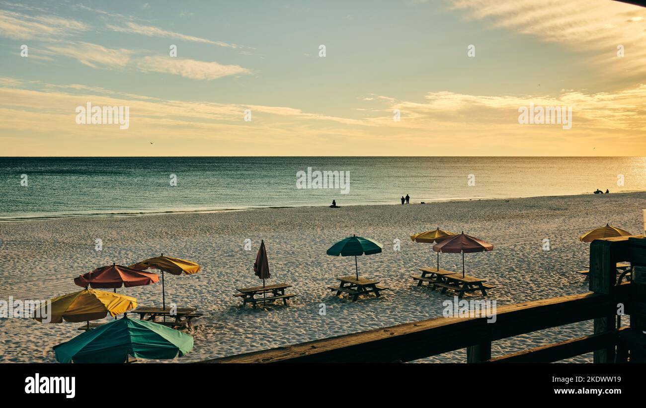 Menschen, die bei Sonnenuntergang an einem Florida Golf Coast Strand in der Nähe von Destin, mit Tischen und Sonnenschirmen, in Florida USA, spazieren gehen. Stockfoto