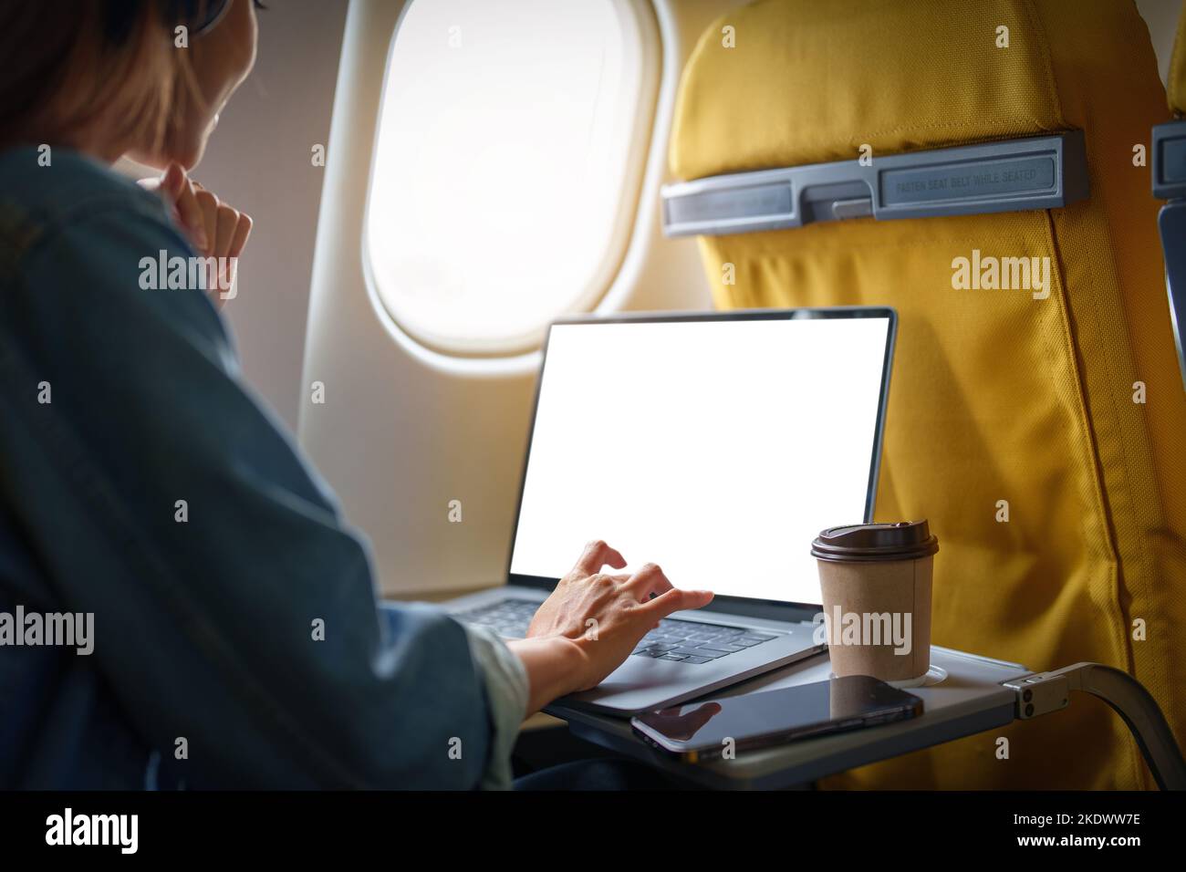 Eine asiatische Frau sitzt in der Economy-Klasse auf einem Fensterplatz und nutzt einen Laptop mit weißem Bildschirm, der während des Fluges Text oder Werbung verwenden kann. Reisen Stockfoto