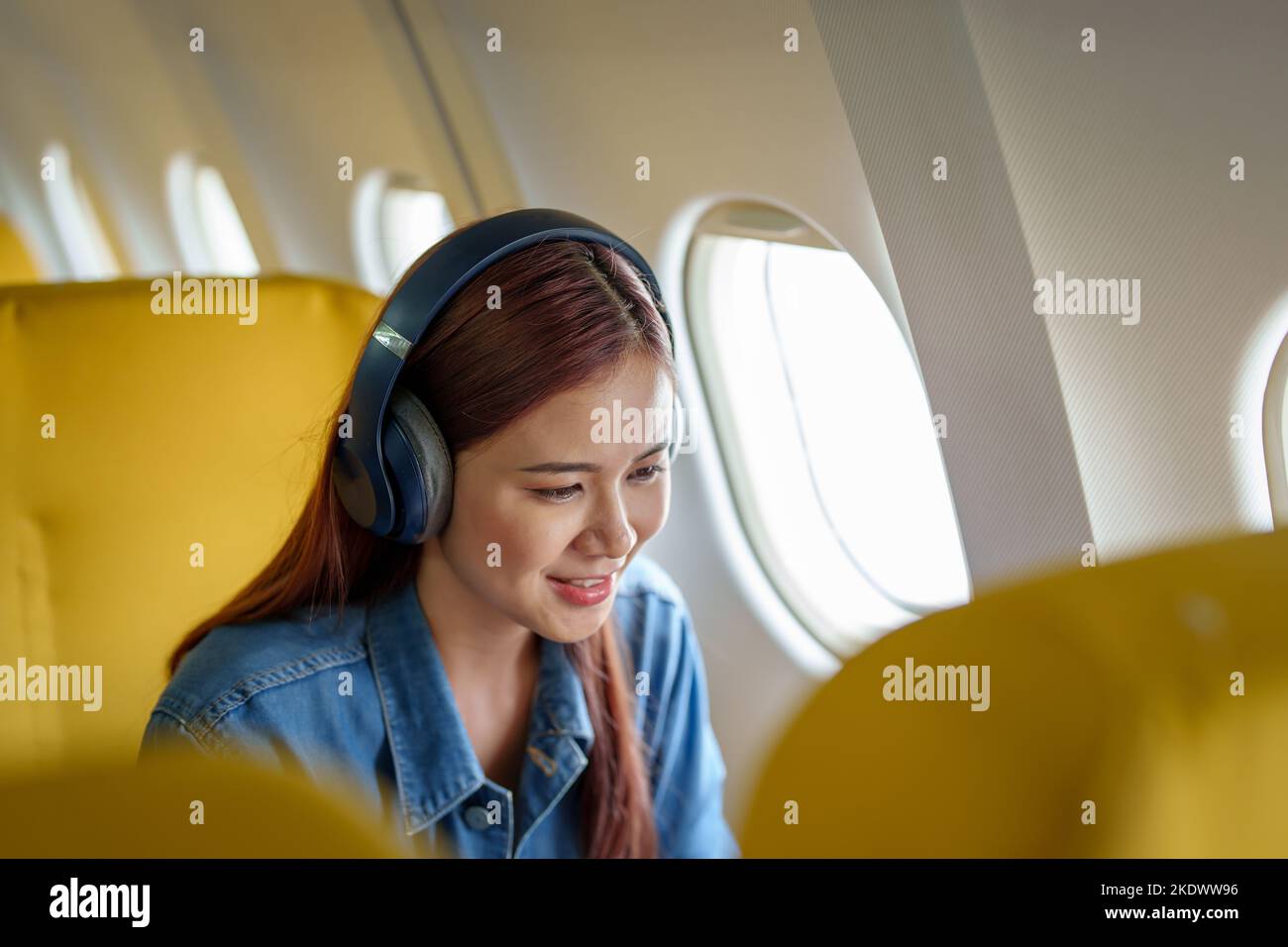 Attraktives Porträt einer asiatischen Frau, die in der Economy-Klasse auf einem Fensterplatz sitzt und während eines Fluges in einem Reisekonzept Musik hört Stockfoto