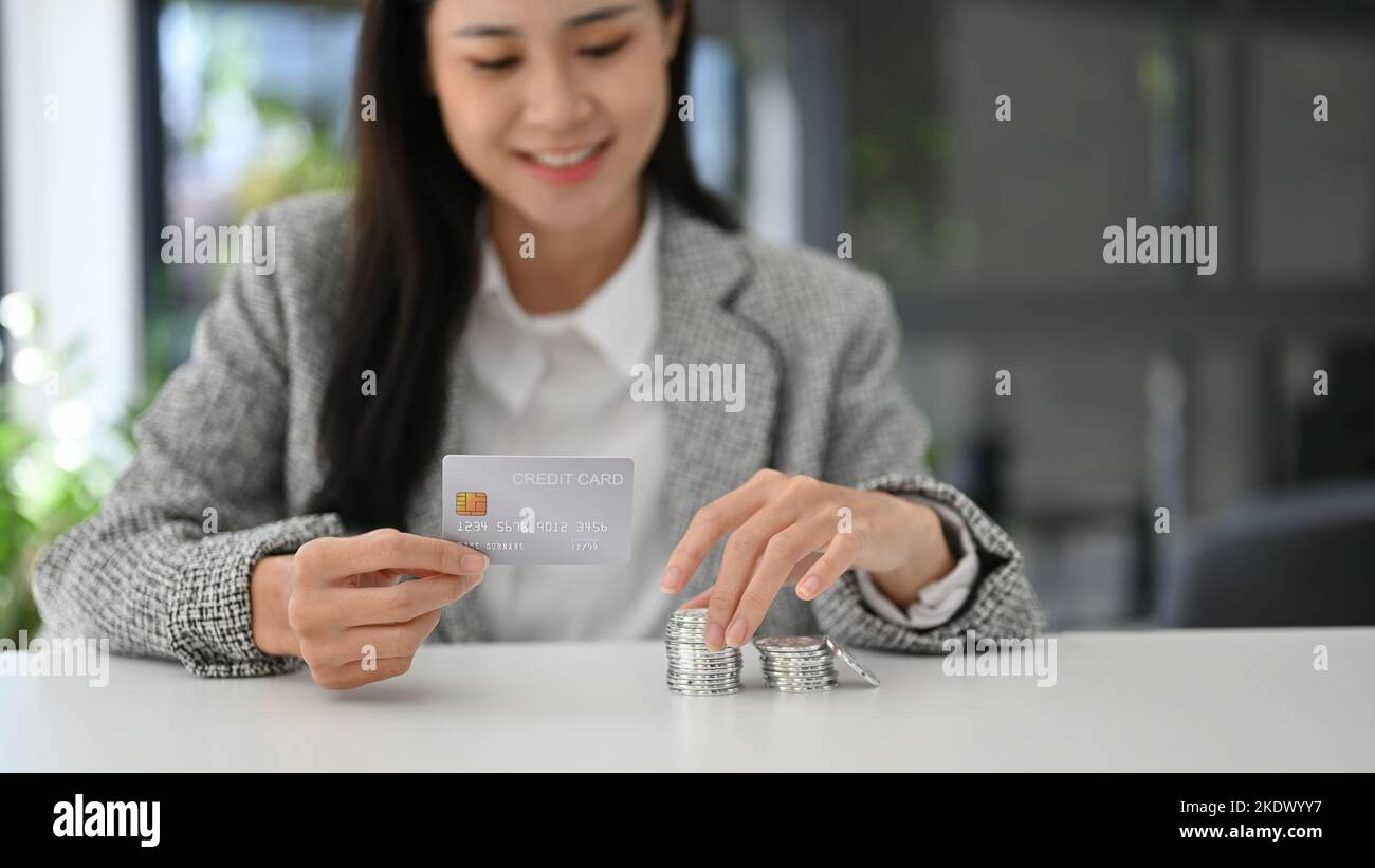 Attraktive asiatische Frau mit Kreditkarte, Stapel von Silbermünzen auf dem Tisch. Finanzen, Banken, Geld, Währung, Investitionen Stockfoto