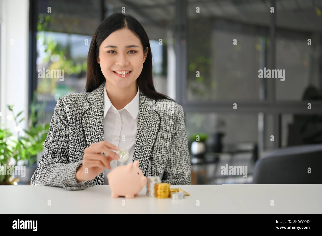 Attraktive und charmante junge asiatische Geschäftsfrau legen Münzen in einem Pin-Sparschwein auf den Tisch. Geld sparen, Investition, Währungskonzept Stockfoto