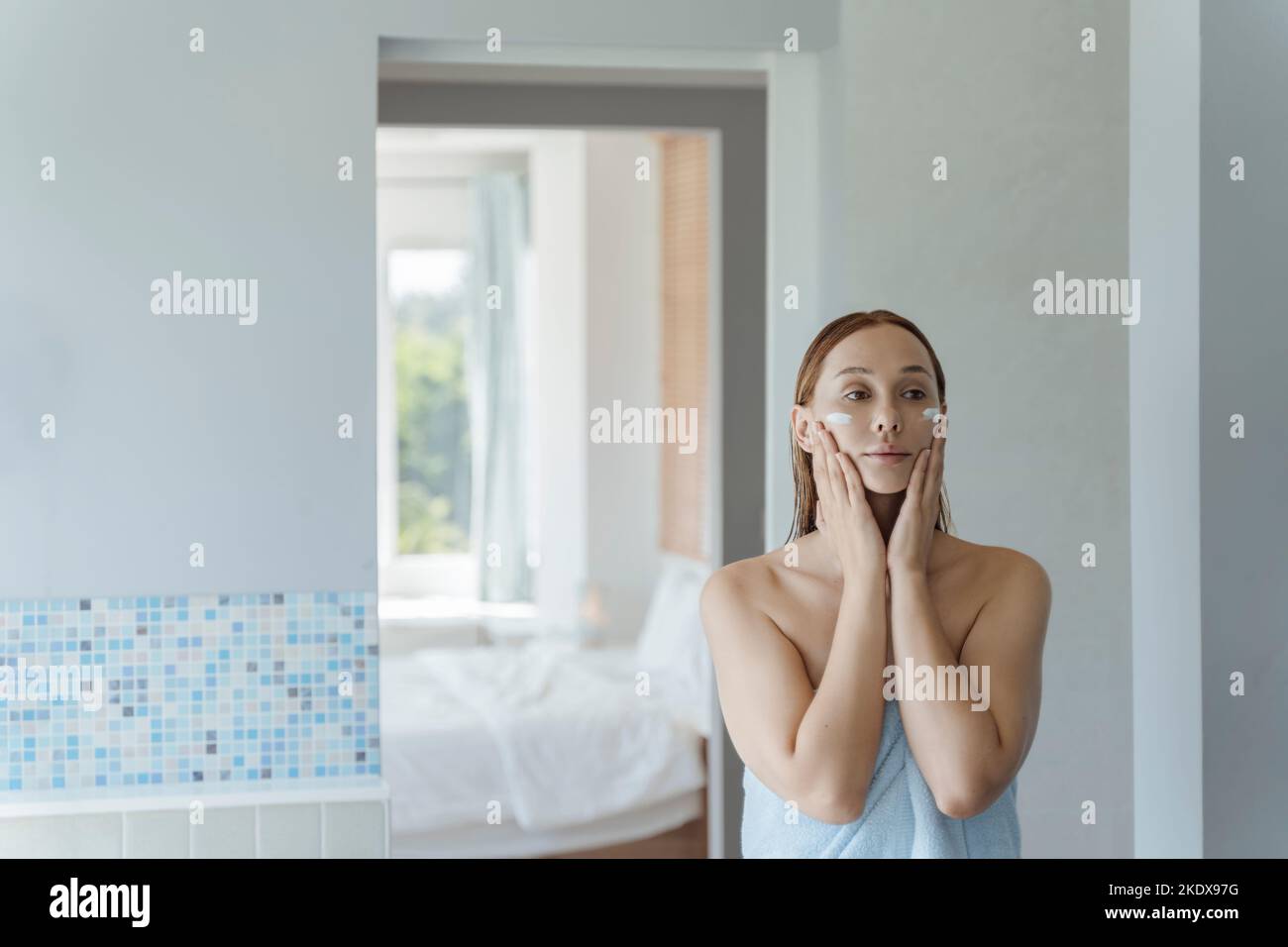Die junge Frau steht nach der Dusche am Spiegel im Badezimmer und gibt ihr Creme auf das Gesicht. Junge Frau verwendet eine Gesichtscreme. Weichfokus. Stockfoto