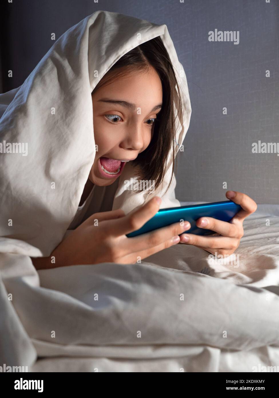 Vertikales Foto eines Teenagers, das nachts mit ihrem Smartphone im Bett unter der Decke spielt. Smartphone- und Gaming-sucht-Konzept. Stockfoto