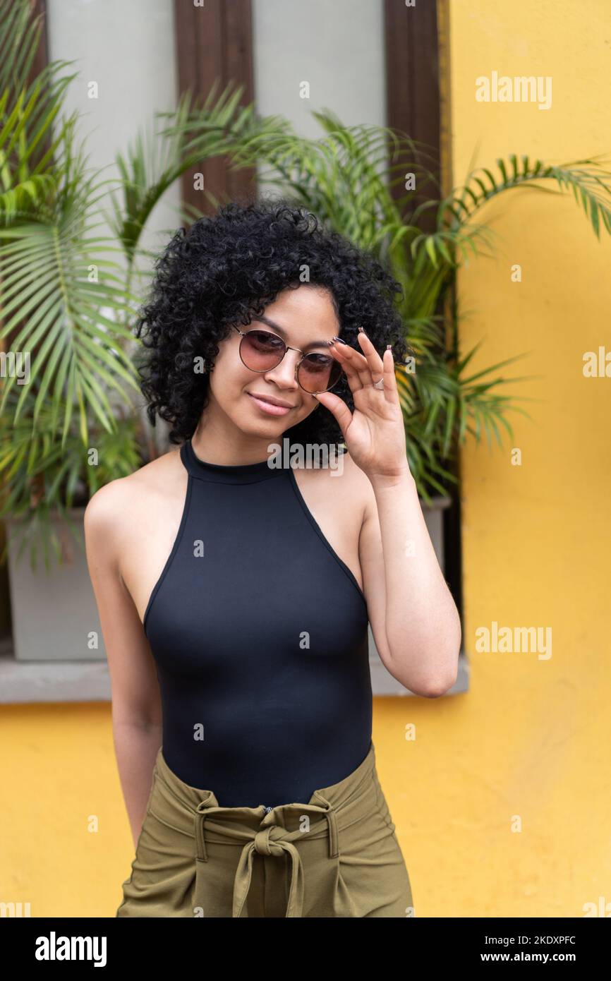Positive hispanische Frau in legerer Kleidung mit lockigem Haar, die trendige Sonnenbrillen anpasst und in die Kamera schaut, während sie in der Nähe des gelben Hauses mit Topf steht Stockfoto