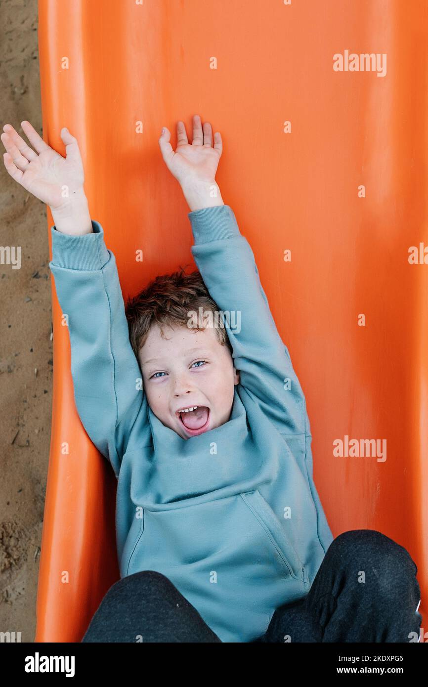 Draufsicht auf einen optimistischen Jungen, der mit einem Lächeln auf die Kamera blickt, während er auf einer orangefarbenen Rutsche über sandigen Boden auf dem Spielplatz spielt Stockfoto
