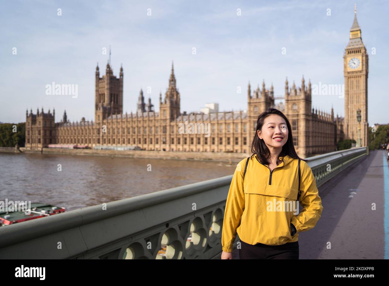 Fröhliche junge asiatische Tourist, die lächelt und weiß wegschaut, steht am Wochenendtag in London auf der Westminster Bridge über der Themse in der Nähe von Big Ben Stockfoto