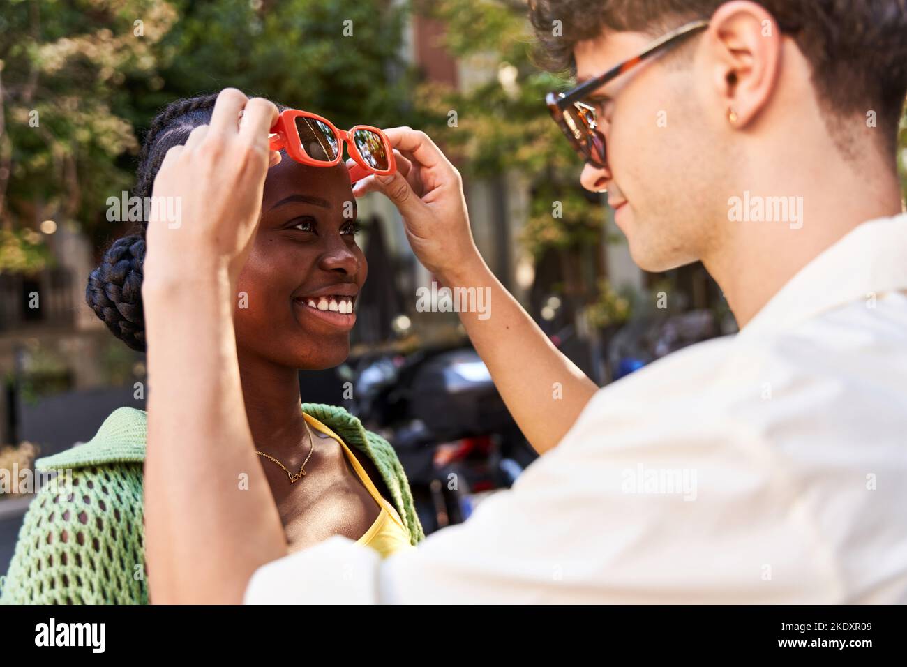 Seitenansicht der Ernte verliebt jungen hispanischen Kerl in weißem Hemd lächelnd und Anpassung Sonnenbrille der fröhlichen afroamerikanischen Freundin auf der Stadt Straße o Stockfoto