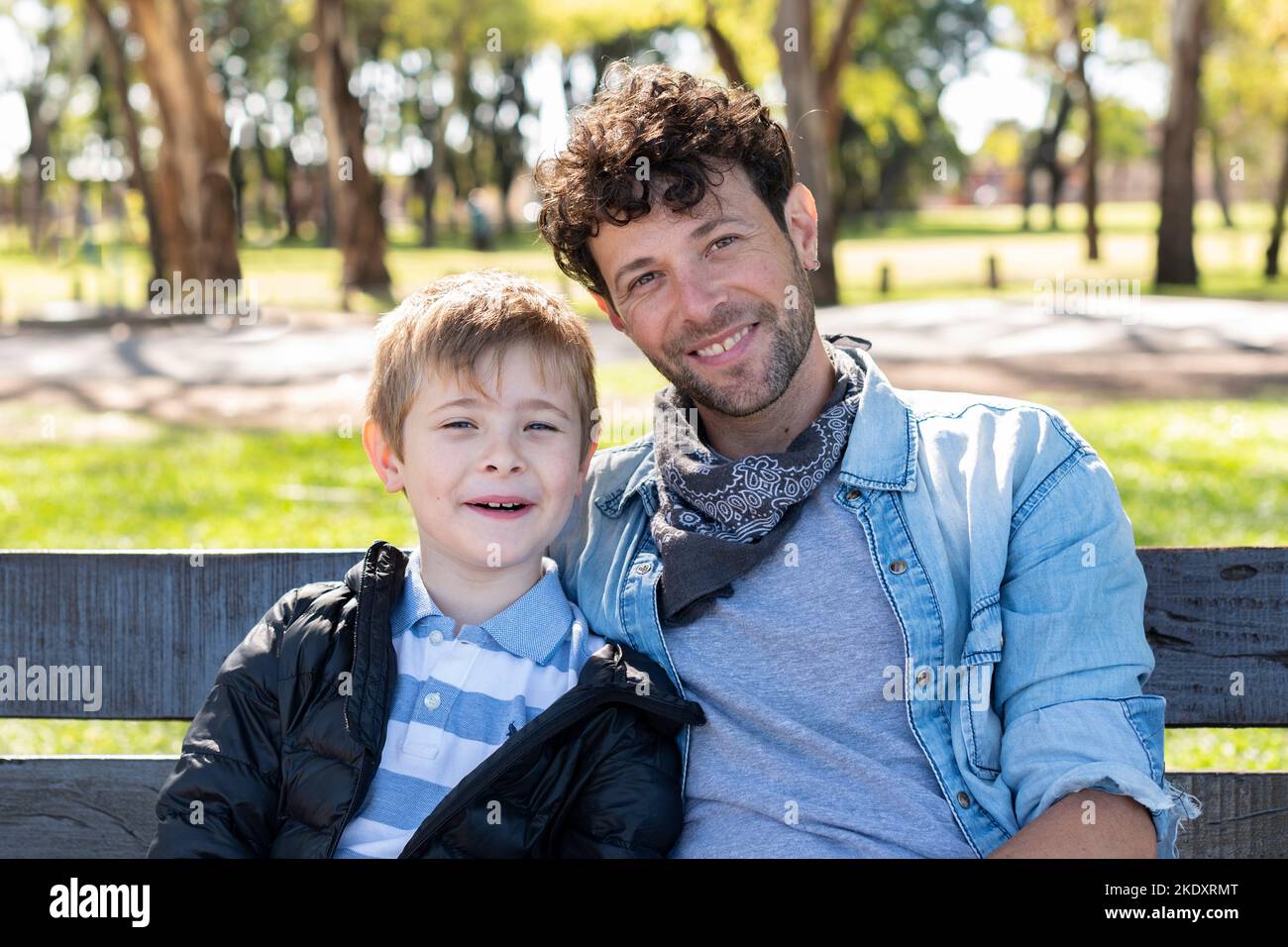 Fröhlicher Vater und Sohn in legerer Kleidung, lächelnd und schauend auf die Kamera, während sie auf der Bank auf unscharfem Hintergrund des sonnenbeschienenen Parks sitzen Stockfoto
