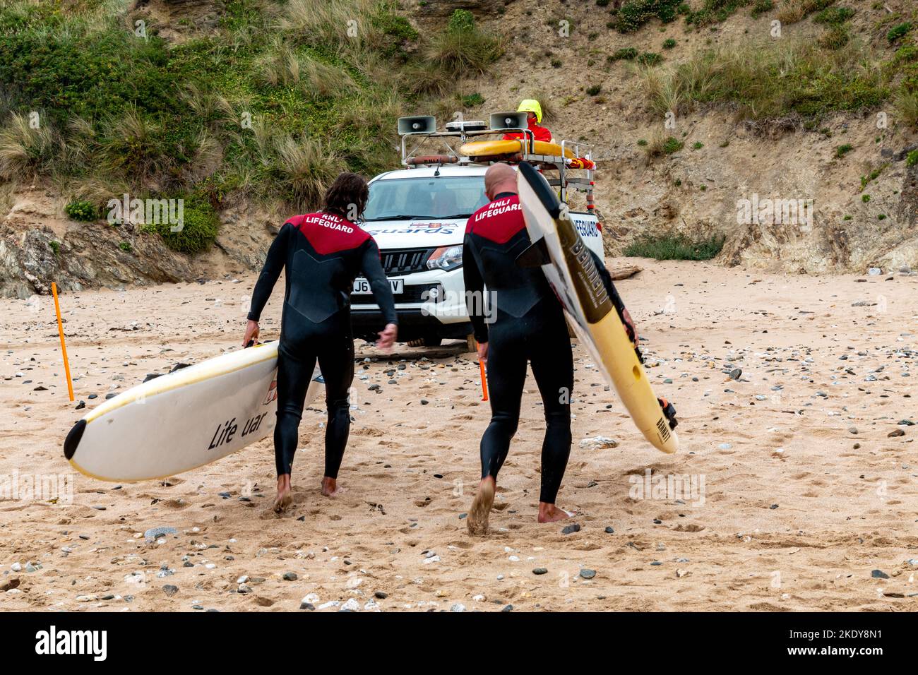 Zwei RNLI-Rettungsschwimmer gehen den Strand hinauf zu ihrem Lastwagen mit Rettungsbrettern, Fistral Beach, Newquay. Stockfoto