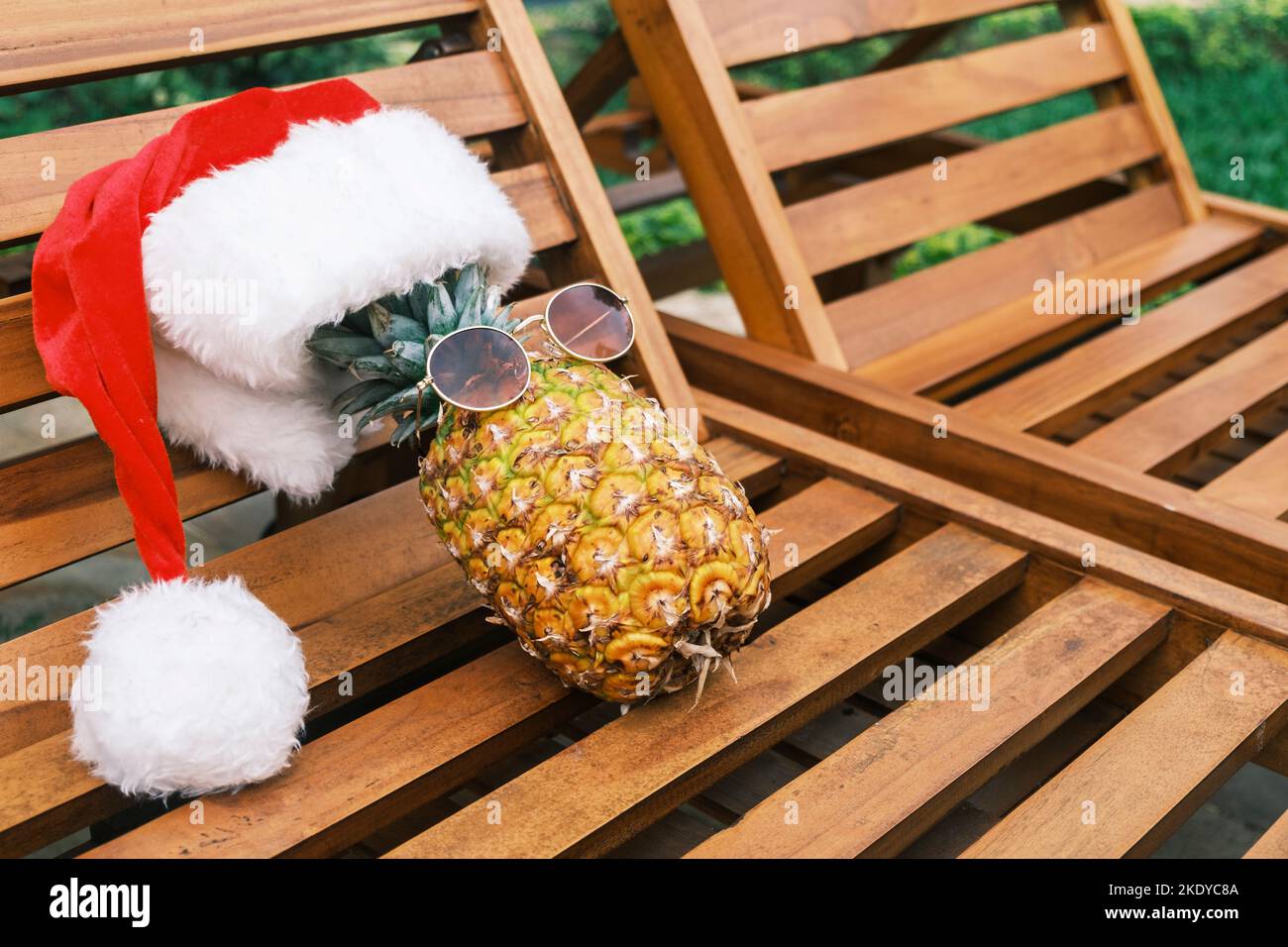 Seitenansicht der entspannenden lustigen Ananas mit Sonnenbrille und Weihnachtsmannhut liegt bei sonnigem Wetter in den Tropen auf einer hölzernen Sonnenliege. Weihnachten in den Tropen Stockfoto