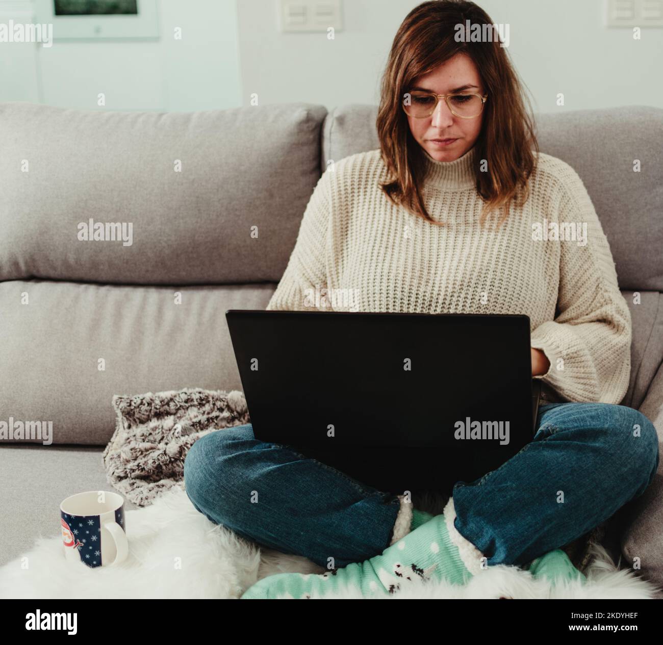 Frau, die ihren Laptop benutzt, um von zu Hause aus zu arbeiten, während sie mit gekreuzten Beinen auf ihrer Couch sitzt Stockfoto