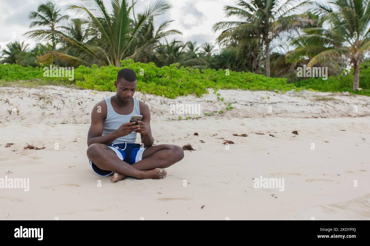 Von vorne sitzt ein schwarzer Reisender auf dem Sand und stöbert während des Sommerurlaubs in den sozialen Netzwerken auf dem Handy. Stockfoto