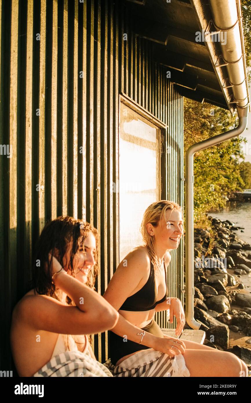 Lächelnde junge Freundinnen, die während des Sonnenuntergangs beim Cottage sitzen Stockfoto