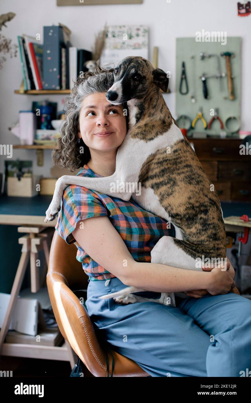 Lächelnde Frau, die Hund umarmt, während sie zu Hause auf einem Stuhl sitzt Stockfoto