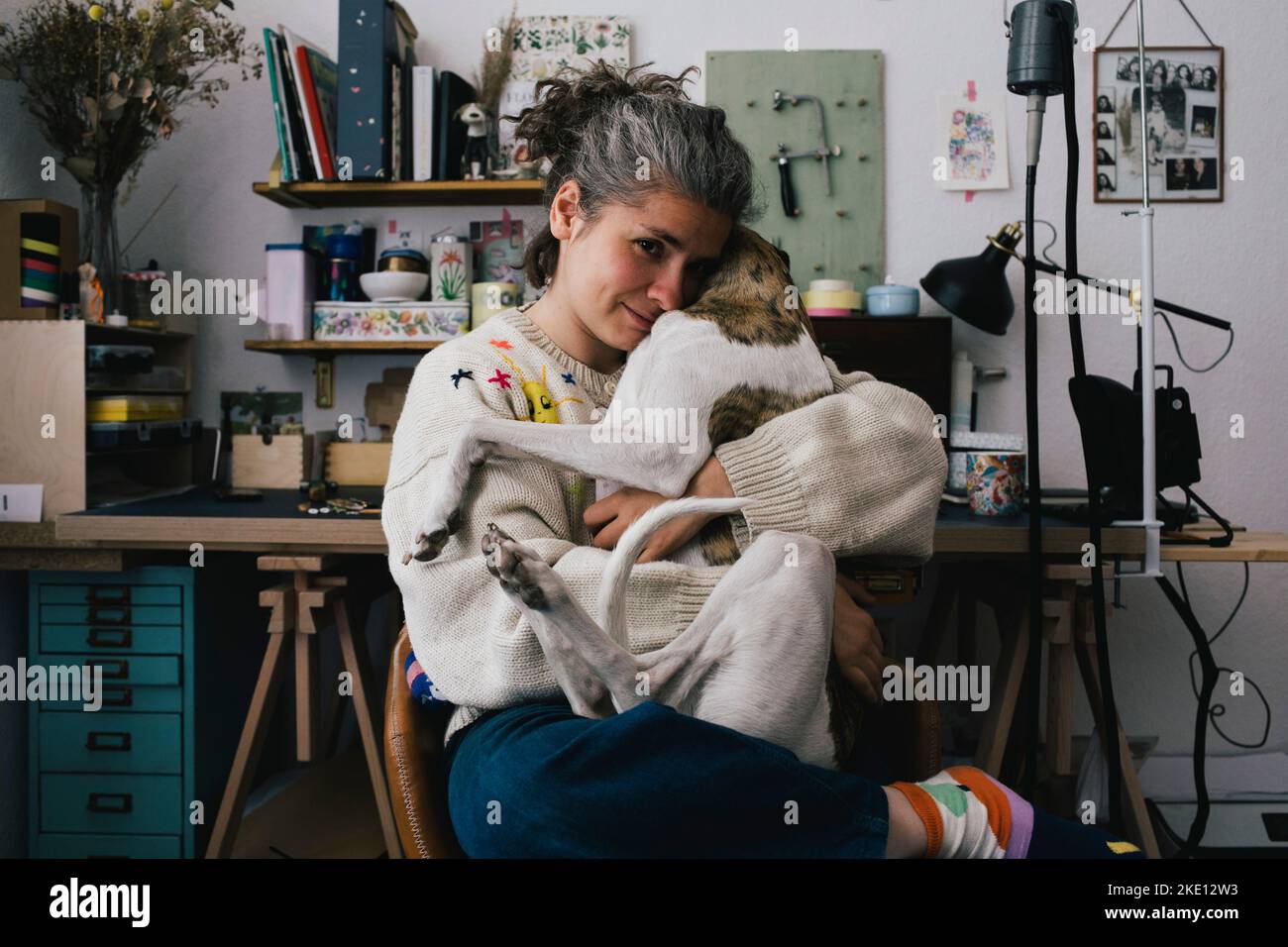 Porträt einer Frau, die einen Pullover trägt und einen Hund umarmt, während sie zu Hause auf einem Stuhl sitzt Stockfoto