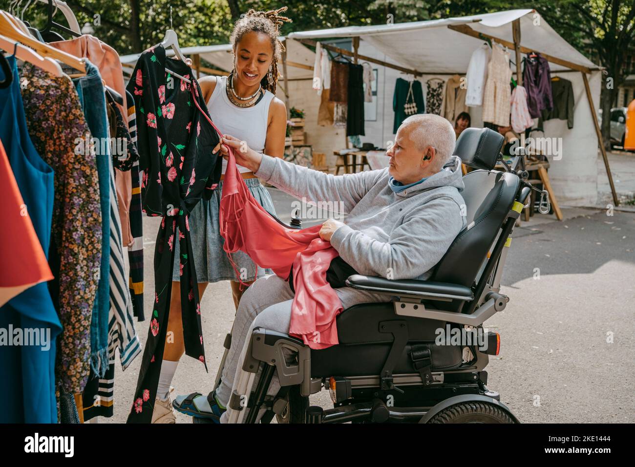 Weibliche Besitzerin, die den Kunden auf dem Rollstuhl beim Kauf von Kleidung auf dem Flohmarkt unterstützt Stockfoto