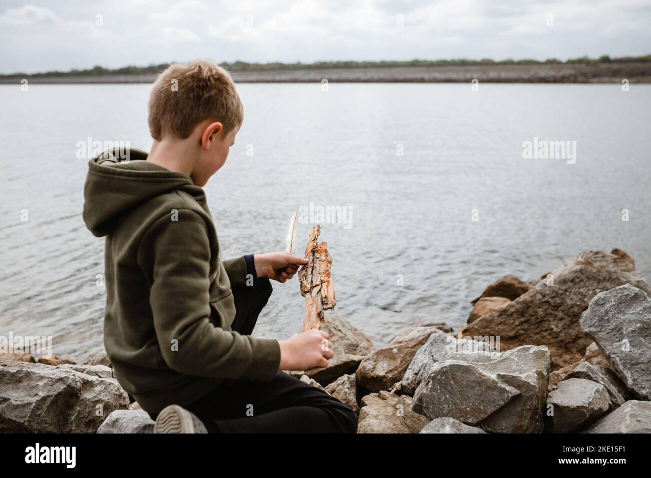 Kleiner Junge, der am See auf Felsen sitzt und Treibholz untersucht Stockfoto