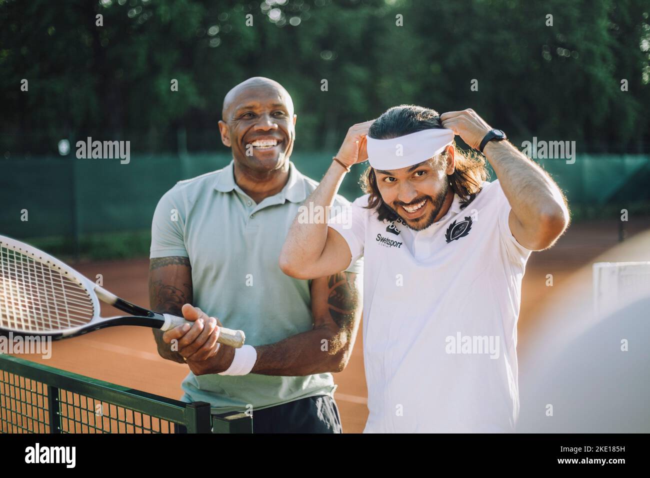 Glücklicher Mann mit Tennisschläger und einem Freund, der das Stirnband auf dem Sportplatz justiert Stockfoto
