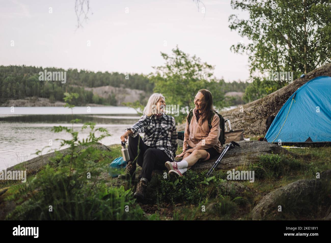 Lächelnde ältere Frauen sprechen miteinander, während sie während des Campens auf Felsen sitzen Stockfoto