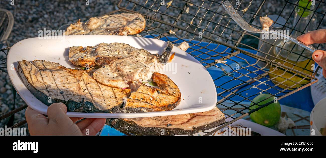 Eine Hand mit einer Gabel, Schwertfischscheiben auf einem weißen Teller und bbq-Grill. Selektiver Fokus auf einem Plastiktisch mit Zitronenfrucht und Garnelenköpfen. Stockfoto