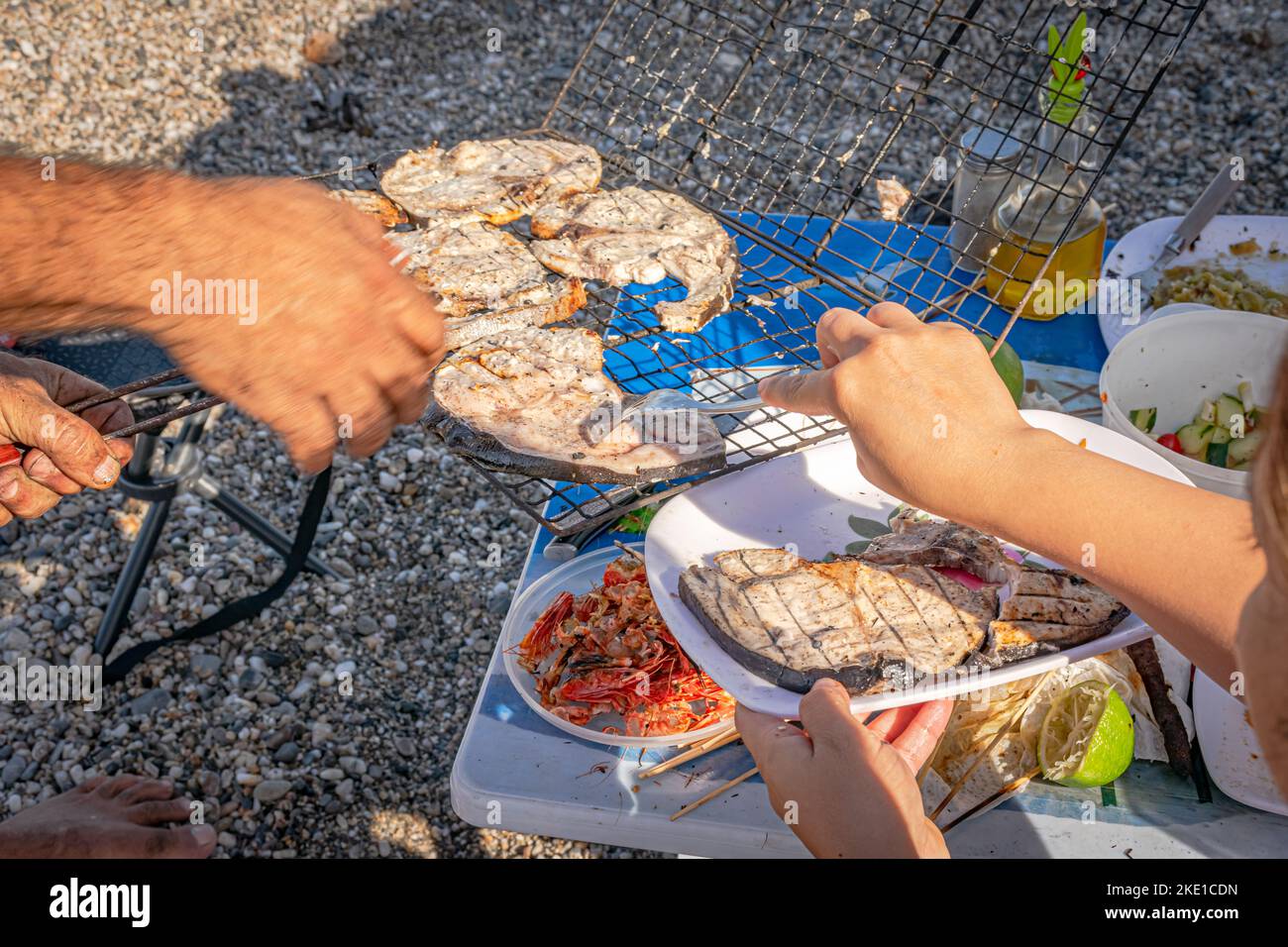 Eine Frau mit einer Gabel, Schwertfischscheiben auf einem weißen Teller und einem grill. Selektiver Fokus auf einem Plastiktisch mit Zitronenfrucht und Garnelenköpfen Stockfoto