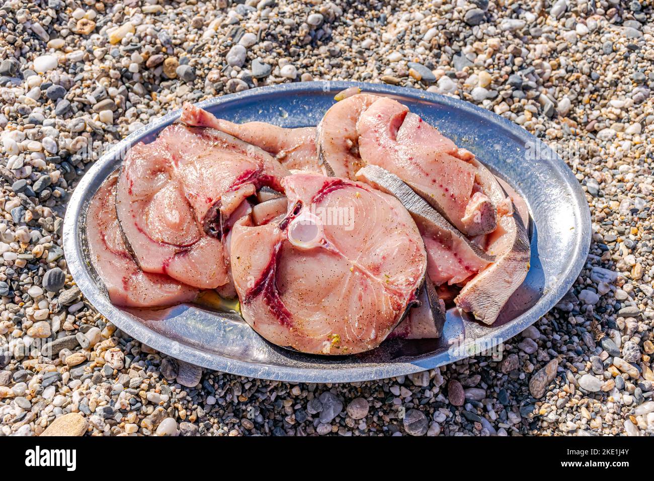 Schwertfischscheiben, die mit Kräutern und Pfeffer gegrillt werden können, auf einer Metallplatte auf dem Boden mit Kieselsteinen am Strand. Direkt über der Außenseite. Stockfoto
