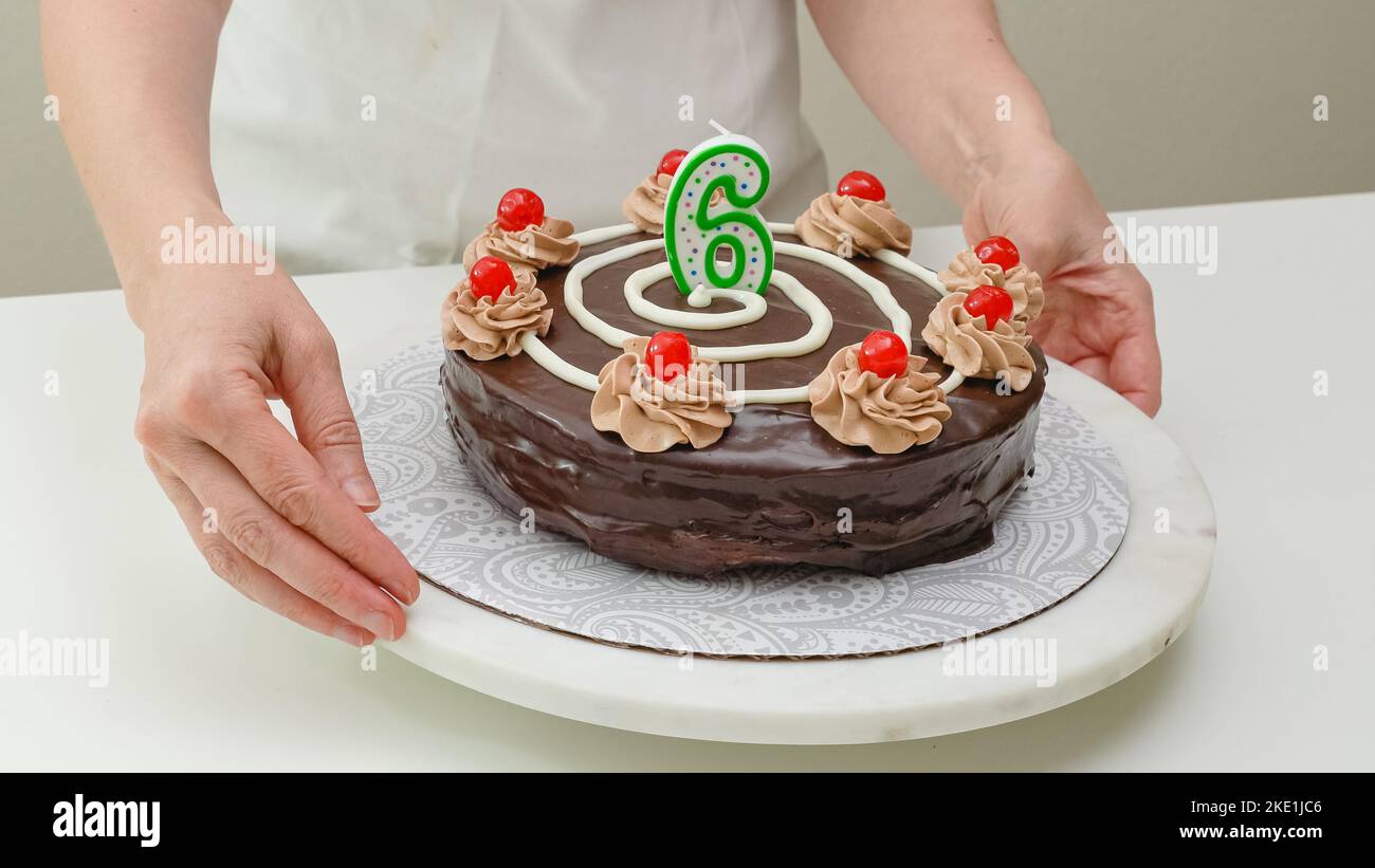 Frau, die Kuchen mit Kerze in Form von Nummer sechs schmückt. Hausgemachter Kuchen mit Schokoladencreme und Kirschen aus nächster Nähe dekoriert Stockfoto