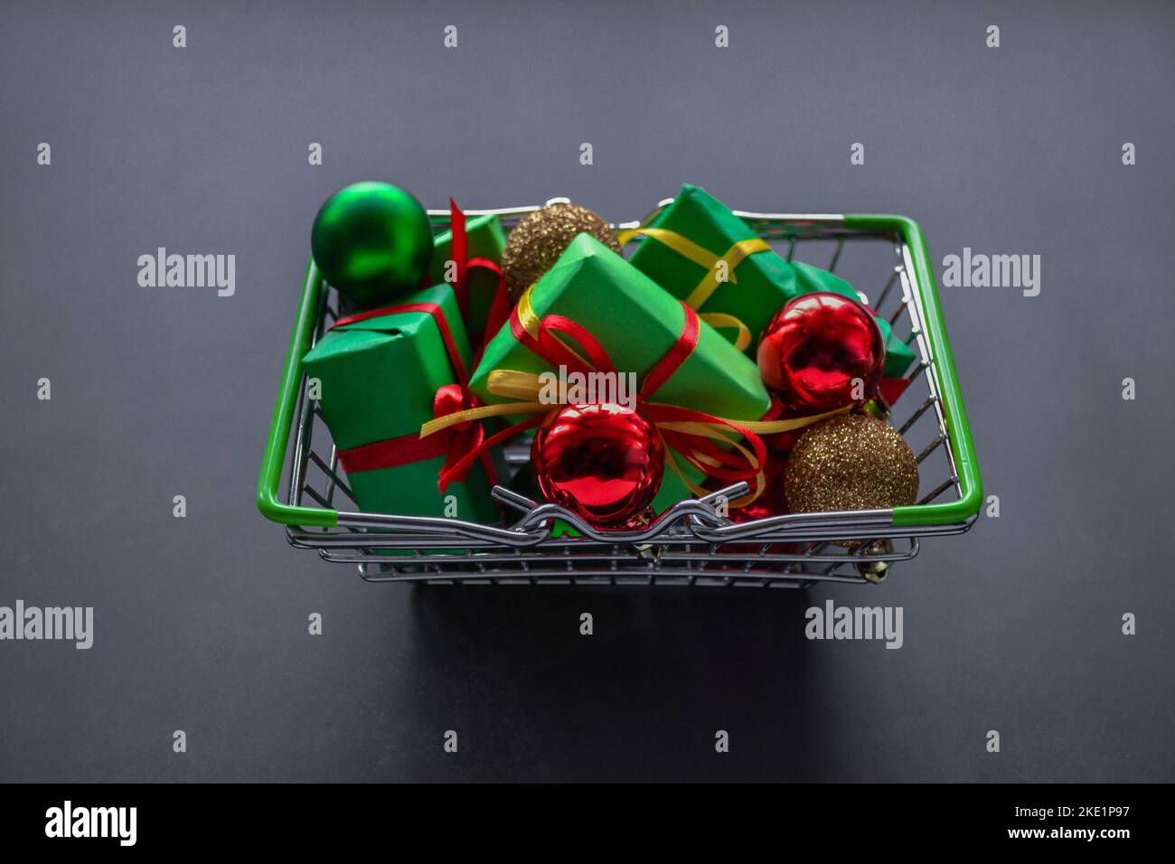 Metall-Einkaufskorb mit Geschenken in grünem Papier mit roten und gelben Schleifen, Weihnachtskugeln aus rot, grün und Gold auf dunklem Hintergrund. Black Friday SA Stockfoto