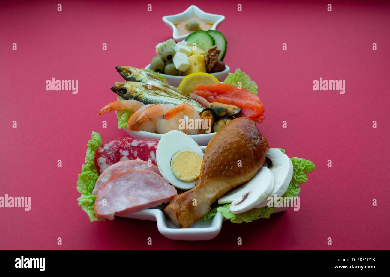 Teller in Form eines Weihnachtsbaums mit Eiweißfutter - Fleisch, Fisch, Käse, Nüsse, etc. Roter Hintergrund. Das Konzept der Keto-Diät behandelt für die holi Stockfoto