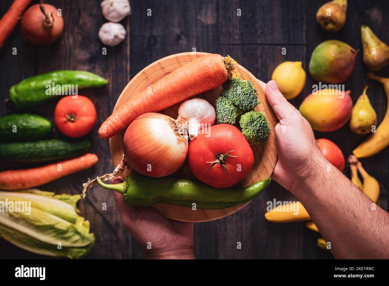 Nahaufnahme der Hände, die einen Teller voller Gemüse auf einem hölzernen Hintergrund mit Obst und Gemüse halten. Gesunde Ernährung und Liebe für frisch und natürlich Stockfoto