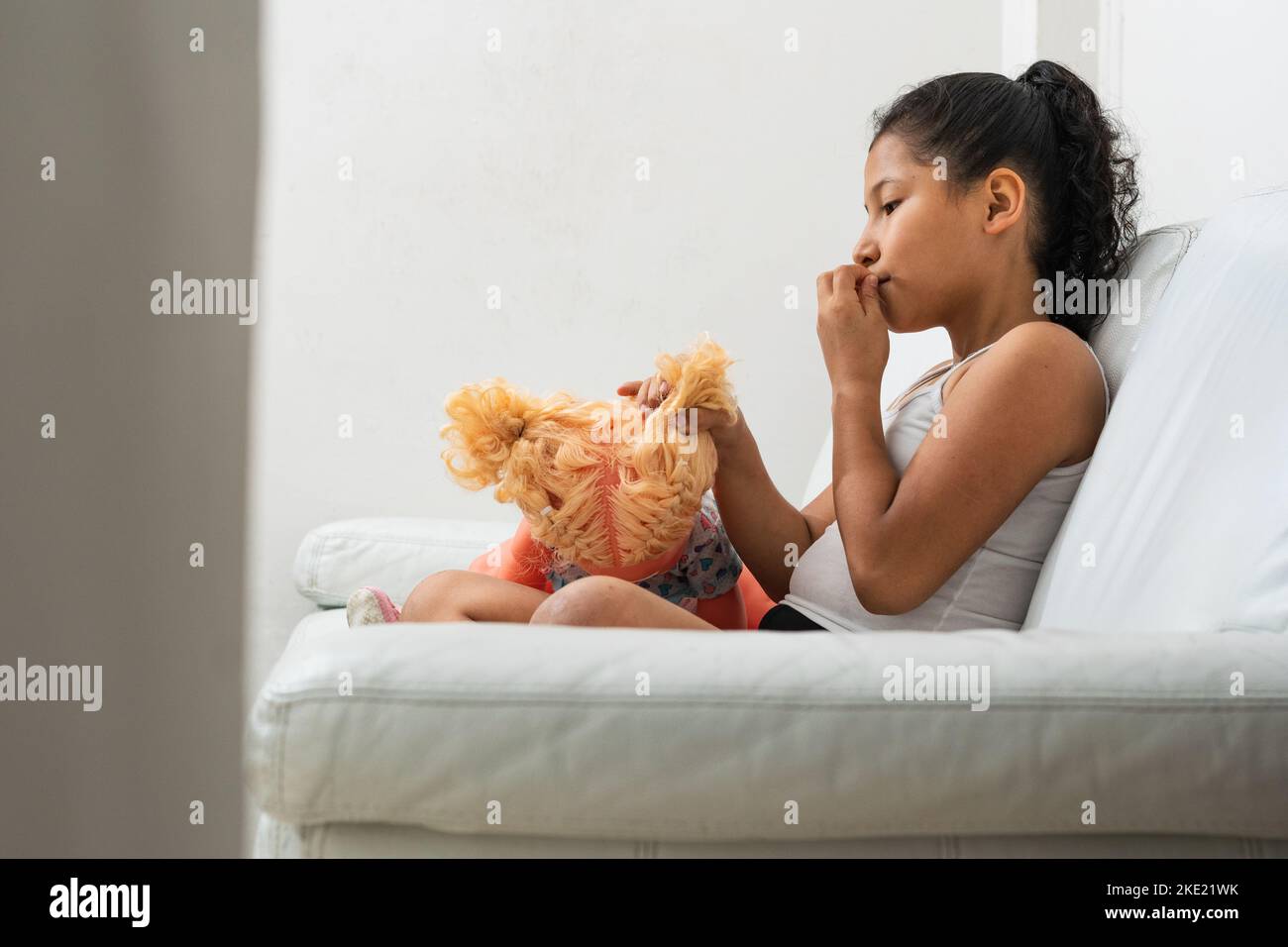 latina Brünette, allein auf dem Sofa sitzend, während sie mit ihrer Puppe spielt. Das Mädchen ist damit beschäftigt, die Haare ihrer Puppe zu kämmen und ein Brötchen in ihren Haaren zu machen. Stockfoto