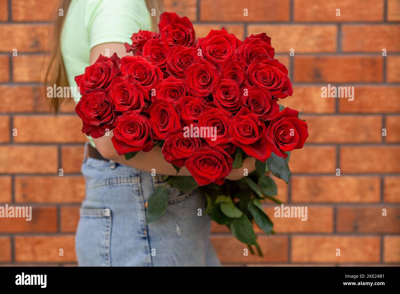 Frau mit einem luxuriösen Strauß frischer roter Rosen vor dem Hintergrund einer Ziegelmauer. Mono Bouquet von roten Rosen in den Händen der Frau. Stockfoto