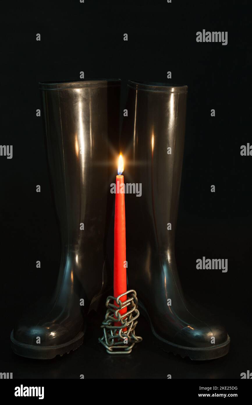Gummistiefel im Kerzenschein. Schwarze Gummistiefel, rote Kerze und Stahlkette, eine ungewöhnliche Weihnachtskarte. Stockfoto