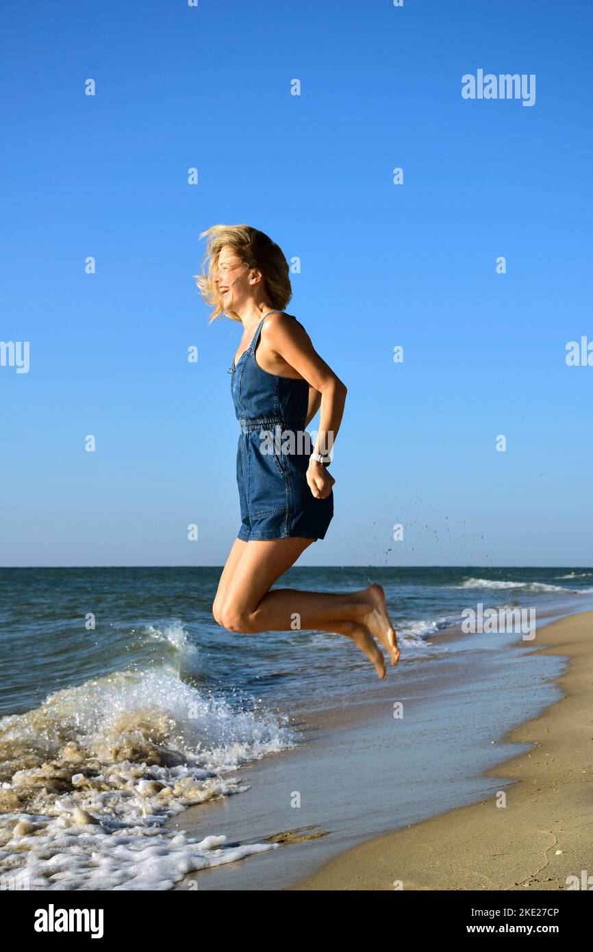Junge, schöne Frau aus weißer Rennblonde springt vor dem Hintergrund der Wellen am Strand und lacht fröhlich Stockfoto