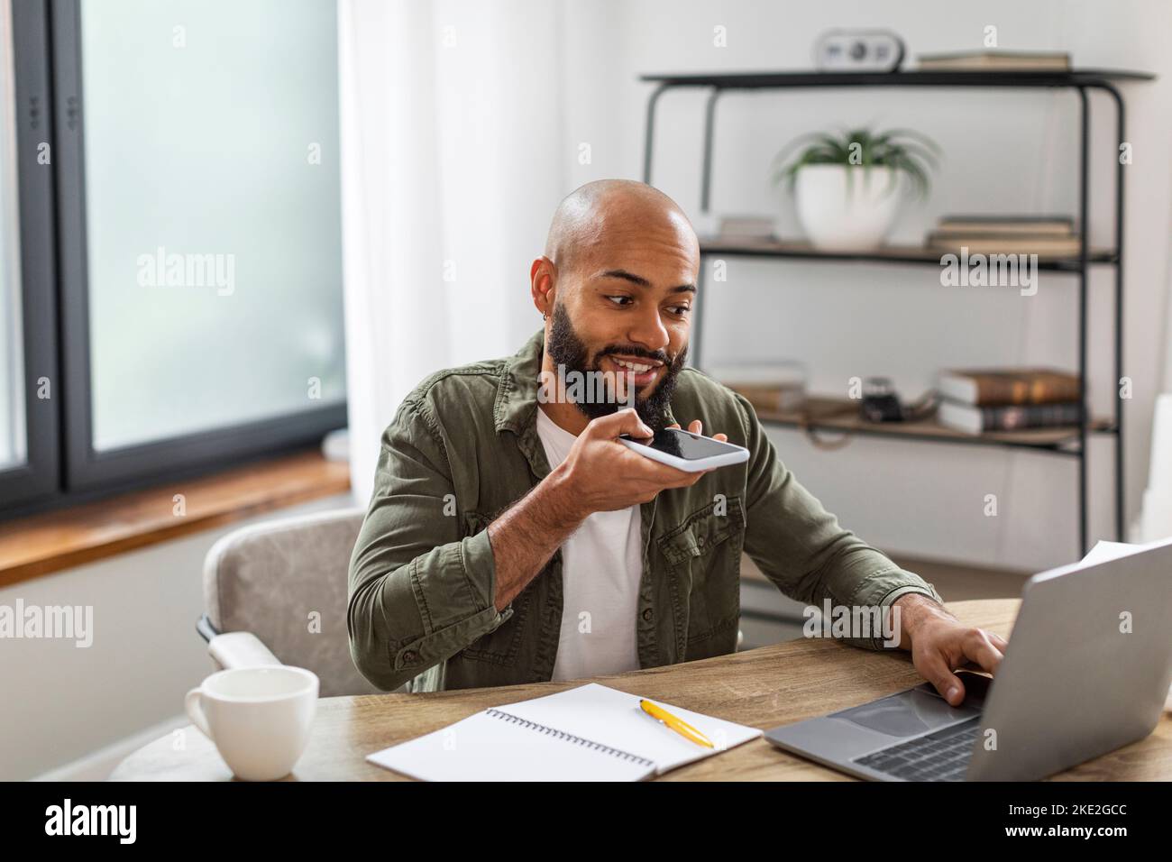 Positiver lateinischer Geschäftsmann, der Audionachrichten auf dem Smartphone und mit einem Laptop aufzeichnet und zu Hause arbeitet Stockfoto