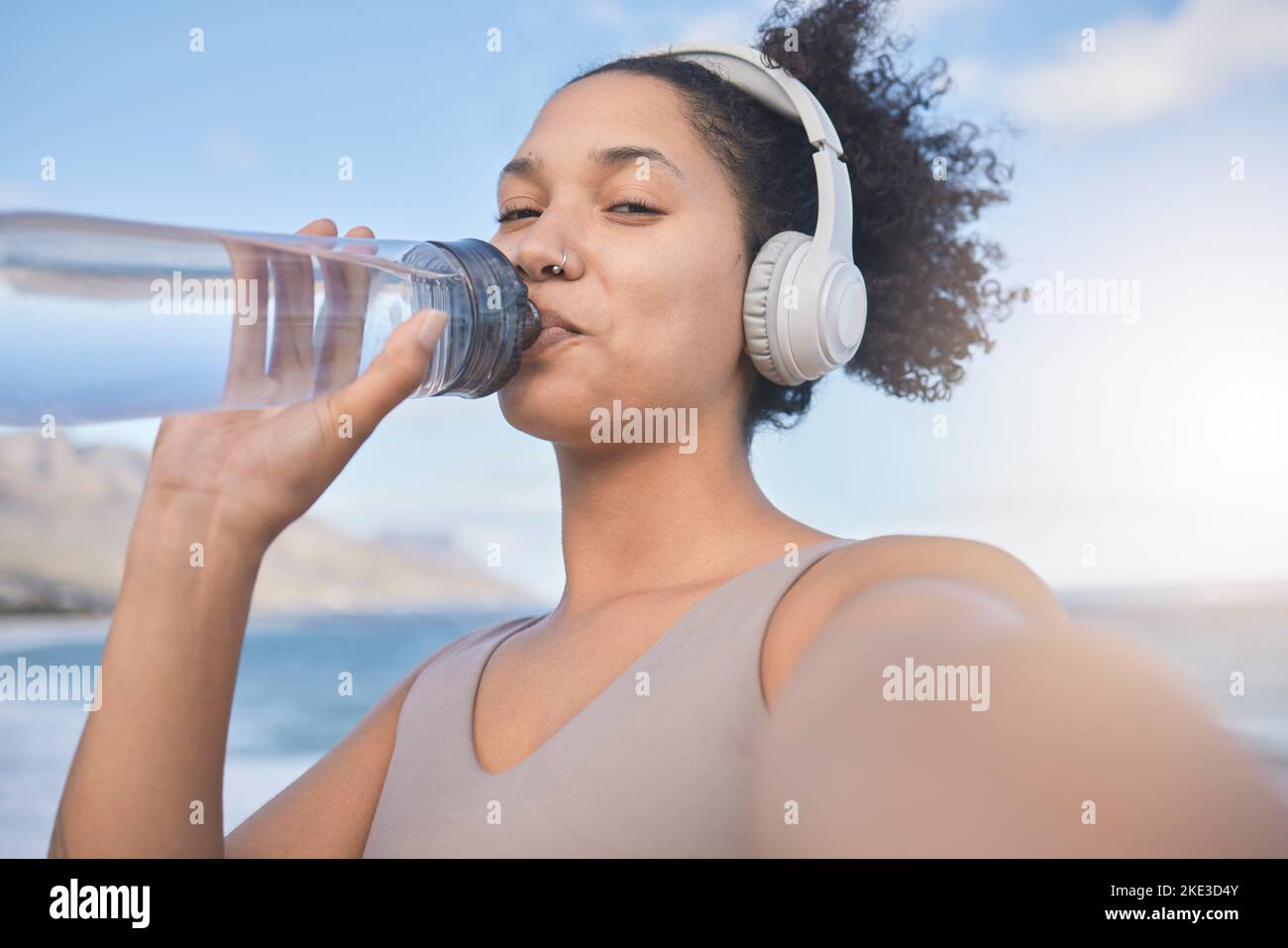 Frau Läuferin, Trinkwasser und Selfie mit Musik, während Sie draußen trainieren, trainieren oder Fitness üben. Schwarze Frau, Kopfhörer und Strand zum Training Stockfoto